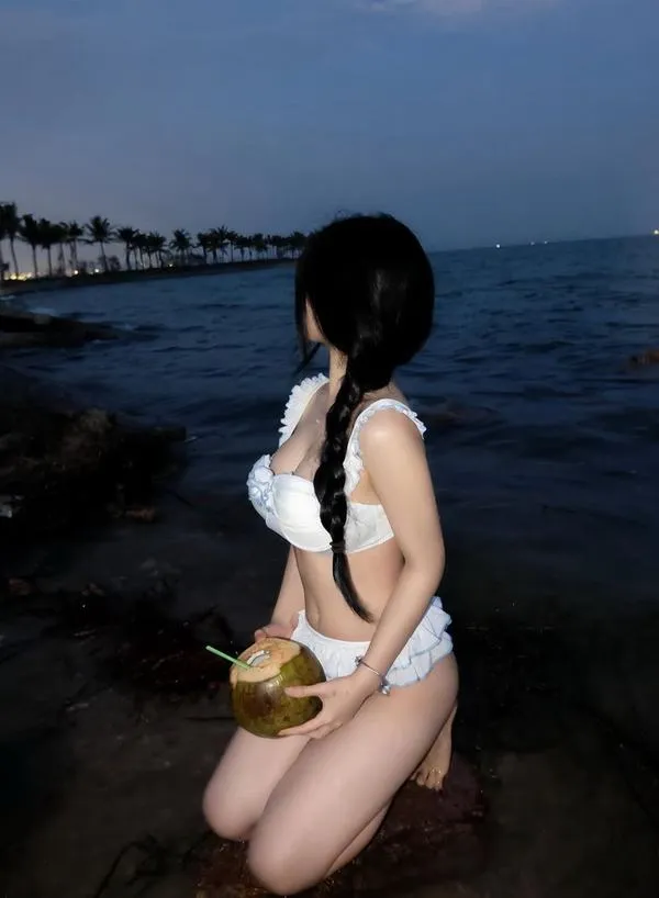 Nàng quỳ bên ghềnh đá, bikini dây ôm dáng, tóc dài phủ lưng, đường cong mềm, mood trầm và nóng.