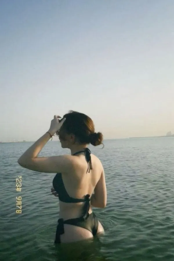 Cô gái đứng giữa mép nước, quay lưng nhẹ, tóc bay lòa xòa, eo thon rõ nét, nền biển xanh yên ả.