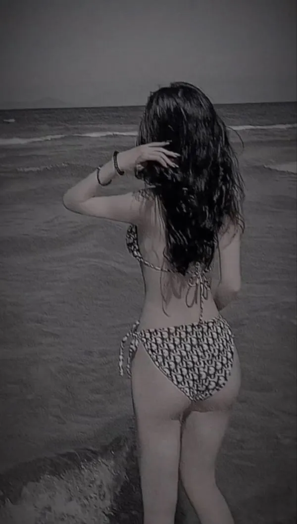 Nàng đứng trên bãi biển vắng, bikini họa tiết, tóc dài ướt nhẹ, lưng cong quyến rũ, cảm giác bí mật.