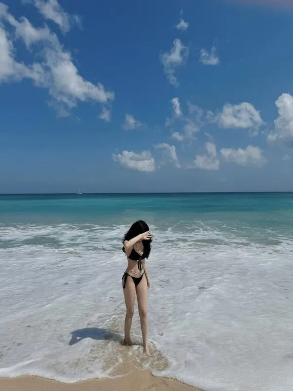 Cô gái nhỏ giữa bãi biển rộng, tay che mặt, bikini gọn gàng, tóc bay, da bắt nắng, dáng đứng mong manh.