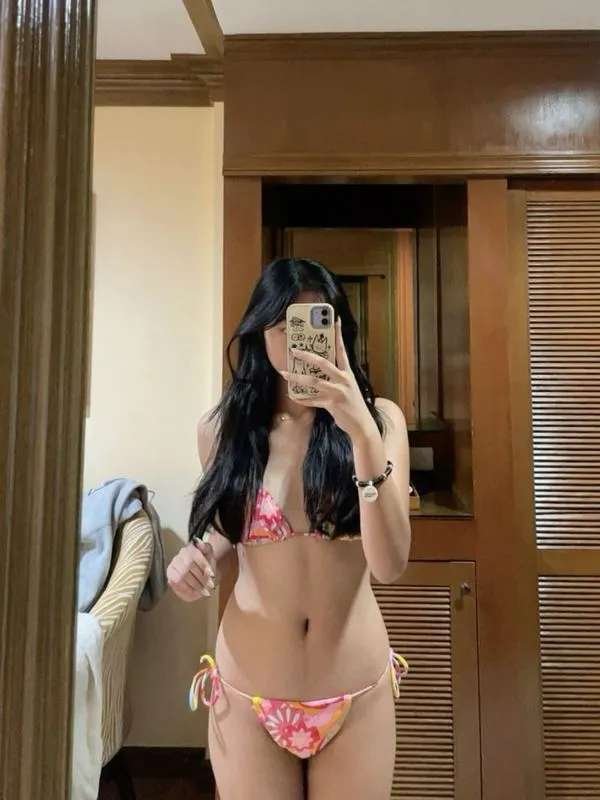 Cô gái đứng trước gương, bikini nhỏ gọn, điện thoại che mặt, da sáng, vòng eo thắt lại, nhìn vừa kín vừa gợi.