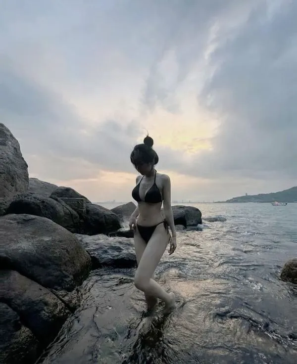 Cô gái đứng trên ghềnh đá, tóc dài bay nhẹ, bikini gọn, da bắt nắng, dáng đứng chắc mà vẫn mềm.