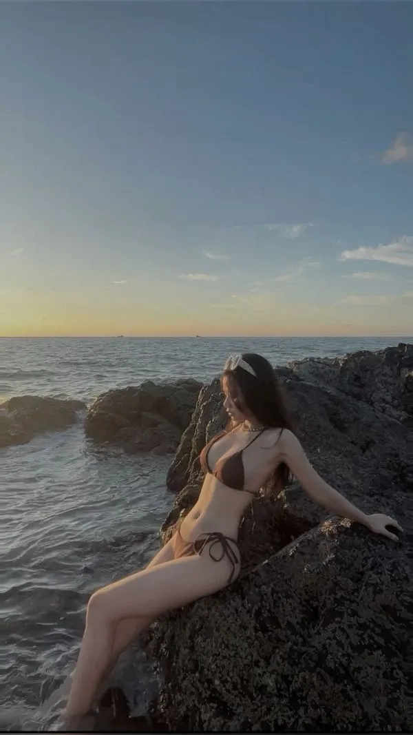 Nàng ngồi tựa đá nhìn biển, bikini ôm eo, tóc xõa, đôi chân thon, ánh nắng khiến da lên màu ấm.