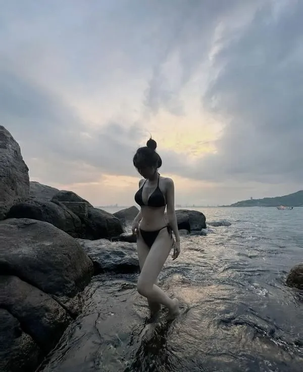 Nàng bước trên ghềnh đá, quay lưng, bikini dây, tóc búi gọn, lưng trần mượt, không khí điện ảnh rõ.