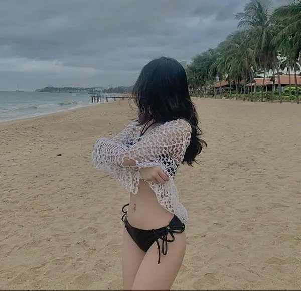 Cô gái đứng trên bãi biển, tay che mặt, bikini tím, tóc xõa, da sáng nổi, dáng mảnh nhìn rất hút.