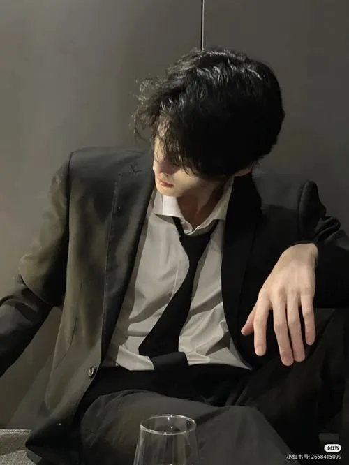 Nam thần lạnh lùng ngồi ghế, outfit tối màu, mắt nhìn xuống như đang giấu tâm sự.