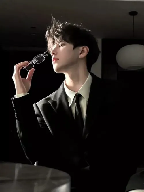 Trai phong trần mặc suit đen, phụ kiện bạc nhẹ, ngồi nghiêng như đang nghĩ chuyện riêng.