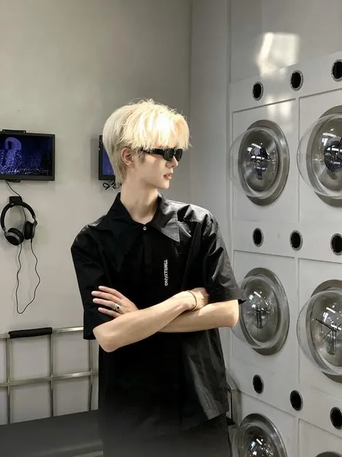 Ảnh trai Douyin tóc vàng đứng trong tiệm giặt, khoanh tay với áo polo đen, góc mặt nghiêng rõ dưới ánh đèn trắng.