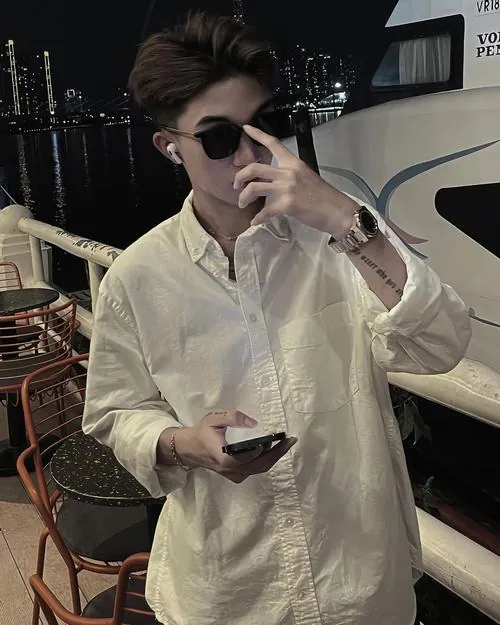 Chàng trai mặc áo sơ mi màu trắng ngà ngồi trong góc tối, đeo kính râm và cầm điện thoại với dáng thả lỏng.
