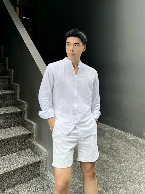 Hình ảnh mỹ nam sơ mi trắng đứng trong nền tối giản, tay bỏ túi, quần trắng đồng màu giúp tổng thể gọn sạch.