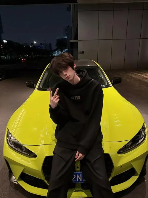 Chàng trai mặc hoodie đen đứng cạnh xe thể thao vàng, tay tạo dáng nhẹ và bối cảnh đường đêm có ánh đèn phản chiếu.