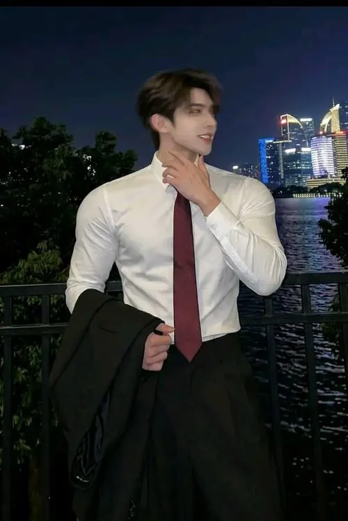 Nam thanh niên sơ mi trắng cà vạt đỏ đứng bên lan can, tay chỉnh cổ áo trong khung cảnh sông nước buổi tối.