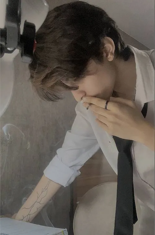 Chàng trai mặc sơ mi trắng cúi đầu sát bàn, mái tóc rũ che một phần gương mặt trong không gian phòng ngủ yên tĩnh.