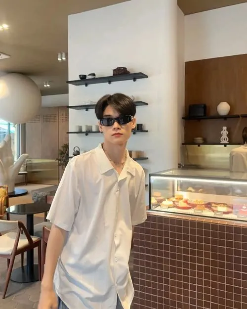 Chàng trai áo sơ mi trắng đứng cạnh máy ảnh phim trong phòng rửa ảnh, khuy áo ngọc trai bắt sáng trên nền tường xám.