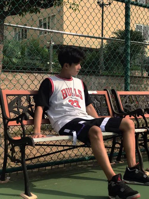 Ảnh trai bóng rổ che mặt ngồi nghỉ trên ghế sân ngoài trời, mặc áo Bulls trắng và quay nghiêng về phía hàng rào.