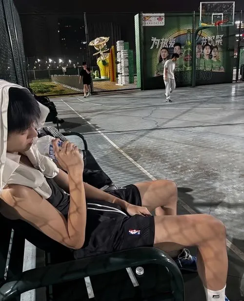 Hình chụp mỹ nam bóng rổ bí ẩn ngồi nghỉ bên sân, tay cầm chai nước và quả bóng đặt gần chân trong ảnh đen trắng.