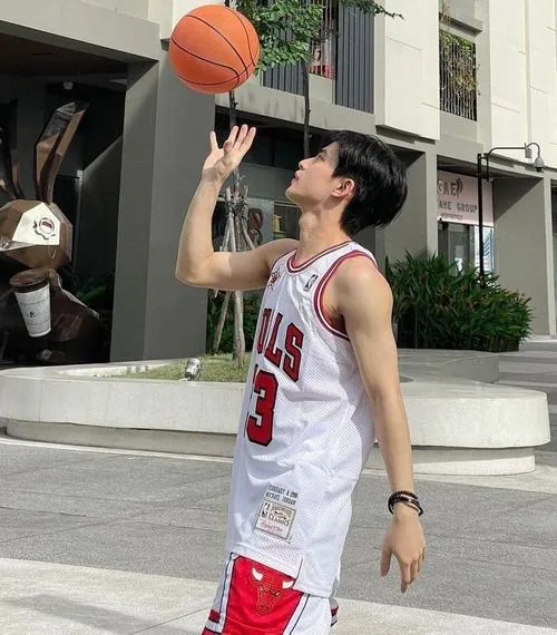 Ảnh góc nghiêng nam thể thao bóng rổ mặc áo Bulls trắng, nâng bóng sát mặt khi đứng trước khu nhà và lan can.