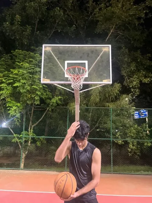 Hình hot boy bóng rổ giấu mặt dùng quả bóng che gần hết khuôn mặt, phía sau là bảng rổ và cây xanh buổi tối.
