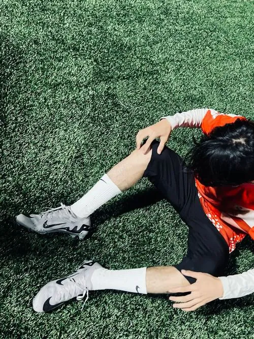 Ảnh trai đá bóng che mặt chụp từ trên cao, chàng trai áo đỏ ngồi co chân trên cỏ, một tay đặt lên tóc sau buổi tập.