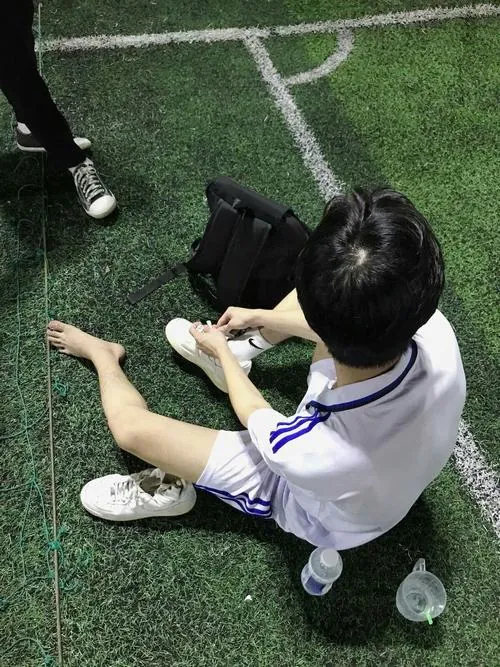 Ảnh hot boy sân cỏ ẩn danh mặc áo trắng viền tím, ngồi xoay lưng và đưa tay lên mặt dưới góc chụp từ cao.