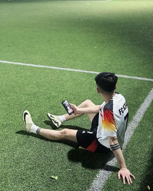 Cầu thủ áo trắng pha cam ngồi duỗi chân, nhìn điện thoại trong tay khi mái tóc che bớt mặt trên nền cỏ xanh.