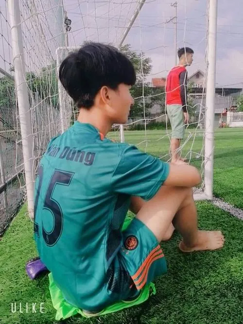 Tựa lưng vào hàng rào, nam sinh áo xanh số 5 ngồi co gối, quay mặt sang bên để lộ mái tóc và bờ vai.