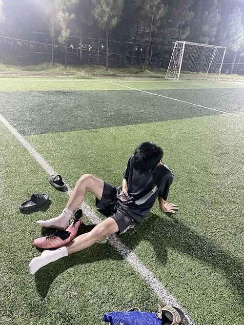 Hình nam sinh đá bóng che mặt cho thấy chàng trai áo đen ngả người trên cỏ, chân duỗi dài cạnh đôi giày tháo rời.