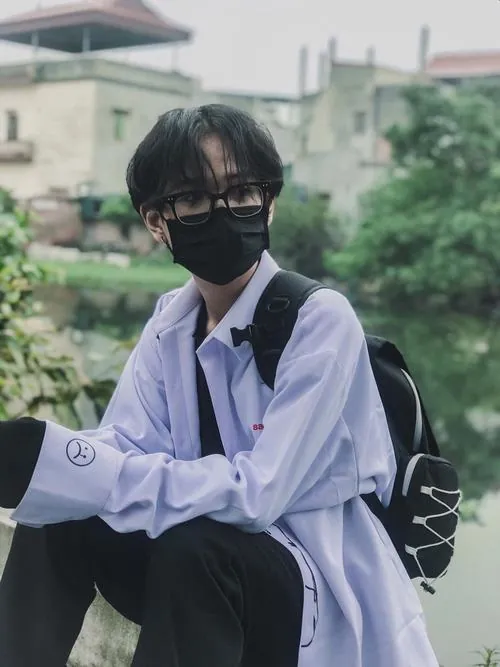 Ảnh soái ca giấu mặt cho thấy chàng trai đeo kính, mặc áo tím nhạt và đứng bên lan can với hàng cây phía sau.