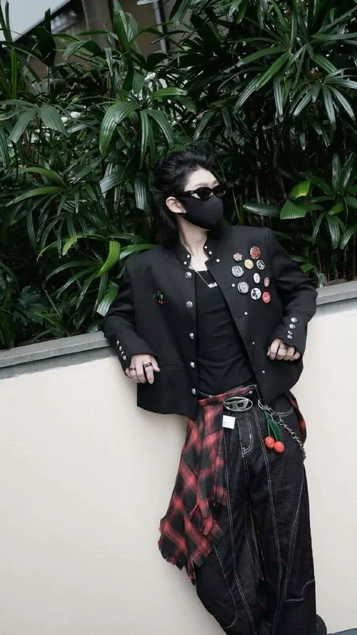 Ảnh hot boy giấu mặt với tóc đen, áo khoác gắn huy hiệu và dáng đứng thư thả bên mảng cây xanh.