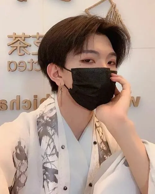 Chàng trai tóc nâu mặc sơ mi sáng họa tiết đen, chống cằm bằng một tay, khẩu trang đen ôm gọn khuôn mặt.