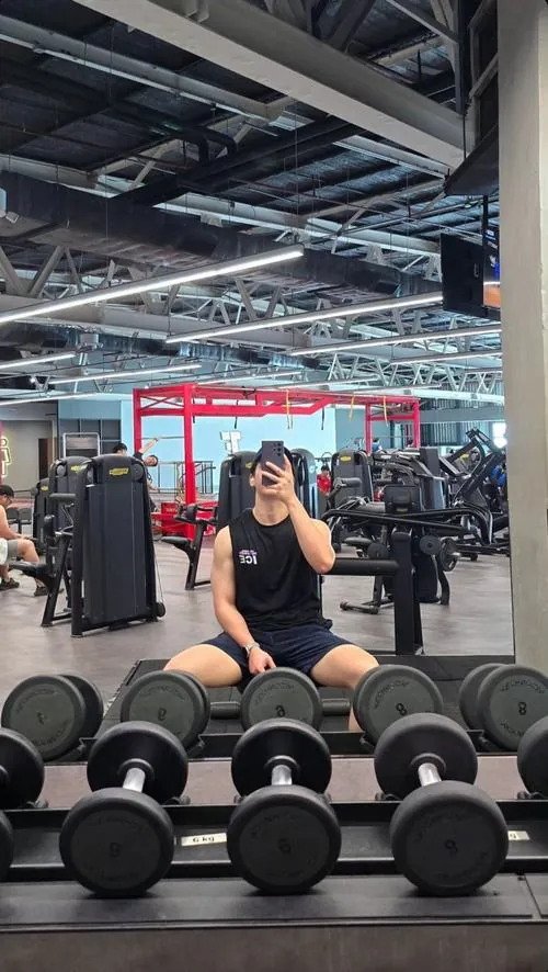 Ảnh trai tập gym đẹp ghi lại chàng trai ngồi trước dãy tạ tay, áo đen, điện thoại che mặt trong phòng tập sáng rộng.