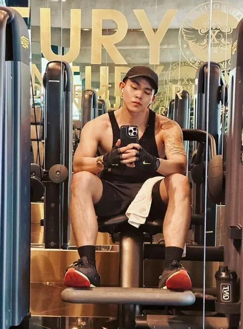 Người đàn ông ngồi trên ghế tập, áo ba lỗ nâu và mũ đen, hai tay đặt gần nhau trong không gian dụng cụ thể hình.