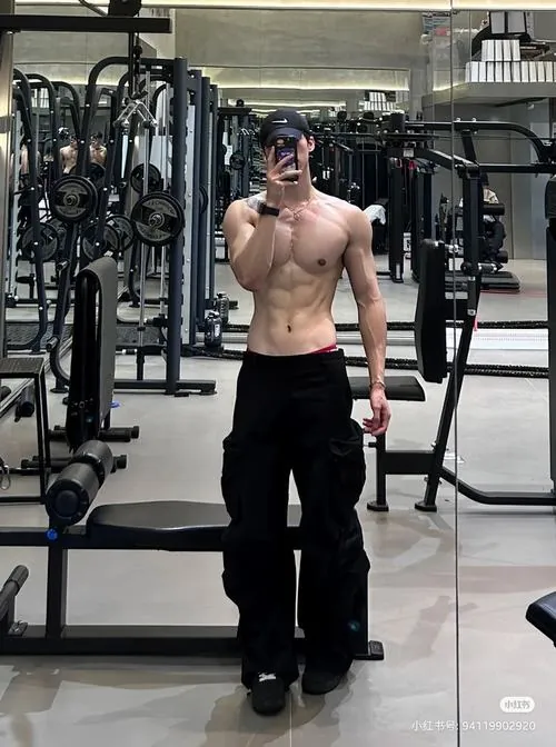 Nam gymer cởi trần bước giữa dãy máy, quần đen đơn giản, điện thoại che mặt và phần ngực nở được ánh sáng nhấn rõ.
