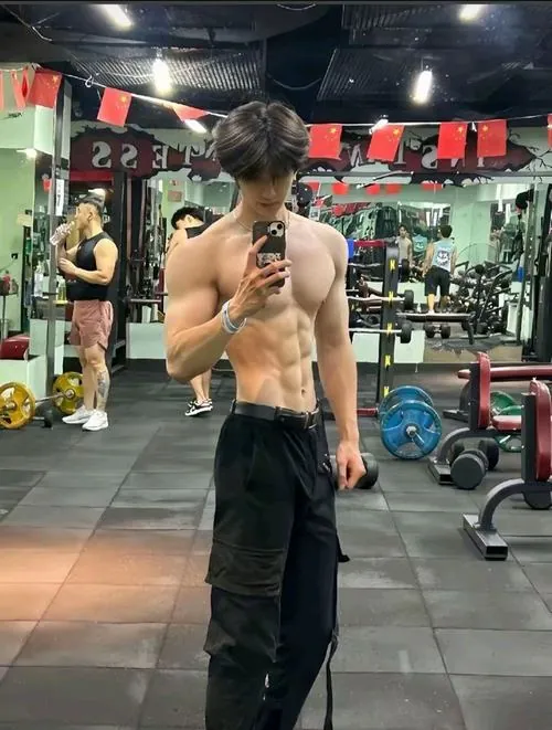 Ảnh trai tập gym đẹp bắt khoảnh khắc chàng trai đứng trong phòng tạ, thân trên để trần, một tay cầm điện thoại trước gương.