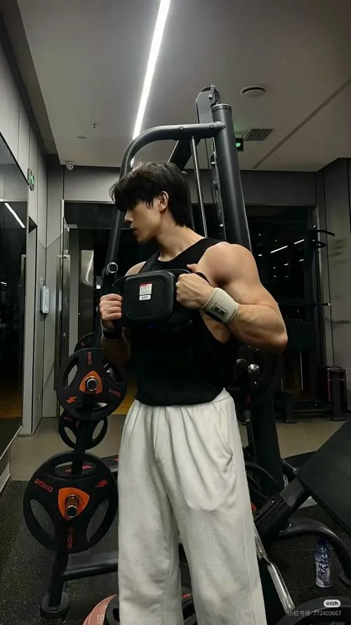 Ảnh trai tập gym đẹp ở góc nghiêng, nam gymer mặc áo sát nách đen, tay cầm điện thoại cạnh máy tập và tạ đĩa.