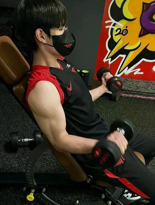 Nam gymer áo đỏ nằm nghiêng trên ghế tập, một tay cầm tạ nhỏ, nền tường vẽ màu tạo cảm giác phòng gym năng động.