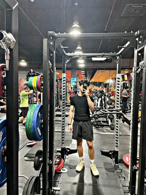 Hình nam gymer vạm vỡ đứng giữa khung rack, hai tay nâng cao, nền sau có đĩa tạ nhiều màu và ánh đèn trắng.