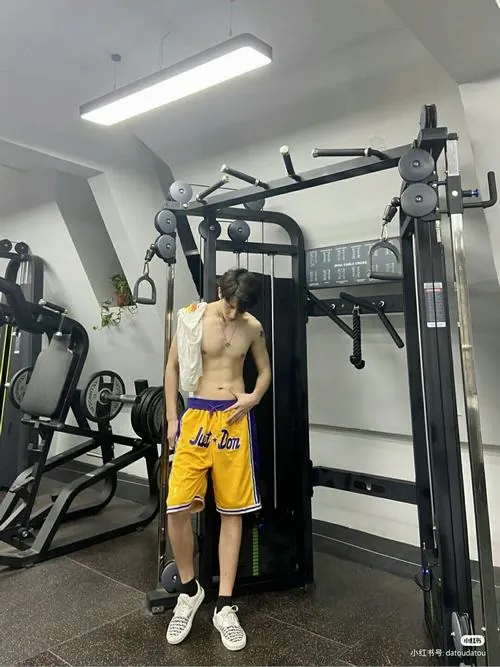 Ảnh nam giới thể thao săn chắc ghi lại chàng trai đứng cạnh máy cáp, quần vàng nổi bật và thân trên để trần rõ nét.