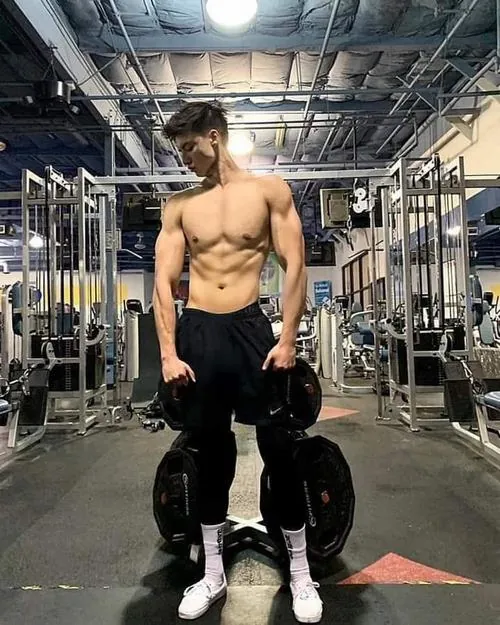 Trong khung hình dọc, nam gymer cầm tạ tay hai bên, thân trên săn chắc nổi bật giữa không gian phòng tập nhiều thiết bị.
