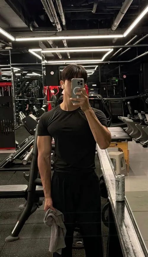 Ảnh trai tập gym đẹp cho thấy người mẫu thể hình mặc áo đen ôm người, tay cầm điện thoại, xung quanh là máy móc ánh sáng dịu.