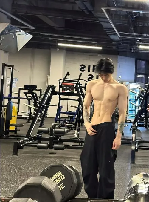 Ảnh trai tập gym đẹp chụp nam gymer đứng thư giãn cạnh máy tập, quần đen rộng, ánh sáng phòng tập phủ đều cơ thể.
