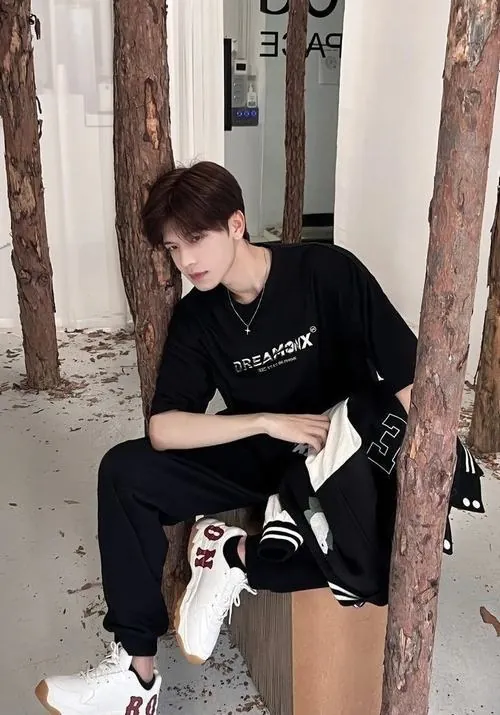 Ngồi tựa bên bậc cửa cũ, chàng trai mặc đồ đen cúi nhìn bàn tay, dáng thả lỏng hòa cùng bức tường sáng màu.