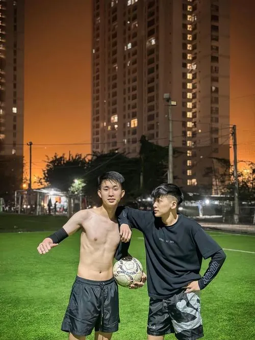 Hai chàng trai đứng trên sân cỏ ban đêm, một người cởi áo cầm bóng còn người kia mặc áo khoác đen.