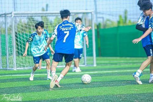 Cầu thủ nam đội mũ len tập kiểm soát bóng trên mặt sân ướt sáng sớm, sương mỏng phủ nhẹ quanh khung thành.
