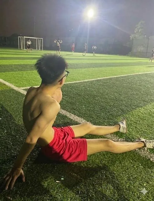 Gương phòng tập phản chiếu cầu thủ mặc áo khoác kéo khóa cao, điện thoại che mắt và đôi găng tay thủ môn đặt cạnh bình nước.