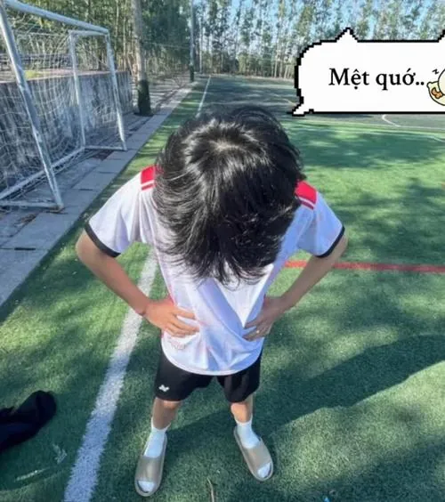 Mưa nhẹ phủ sân bê tông khu phố, chàng trai đội mũ trùm và che miệng bằng cổ tay, giày đinh xách gọn trong túi lưới.