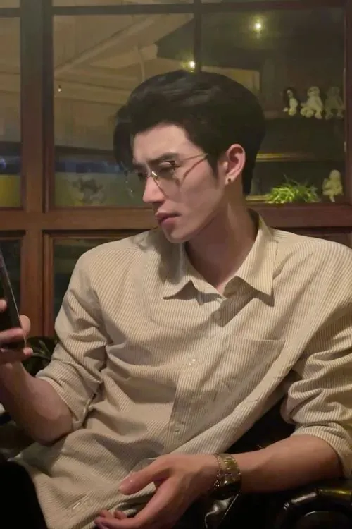 Chàng trai đeo kính mặc sơ mi màu be ngồi bên bàn, ánh nhìn hướng xuống và tư thế thư giãn tạo vẻ trí thức gần gũi.