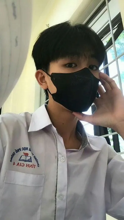 Hình trai đẹp lớp 7 che mặt với khẩu trang đen, mặc sơ mi đồng phục và nghiêng đầu cạnh khung cửa trong phòng.