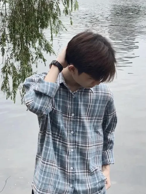 Nam sinh áo sơ mi kẻ xanh đứng bên bờ nước, cúi đầu và đưa tay chỉnh tóc, tạo dáng tự nhiên giữa khung cảnh nhiều cây.