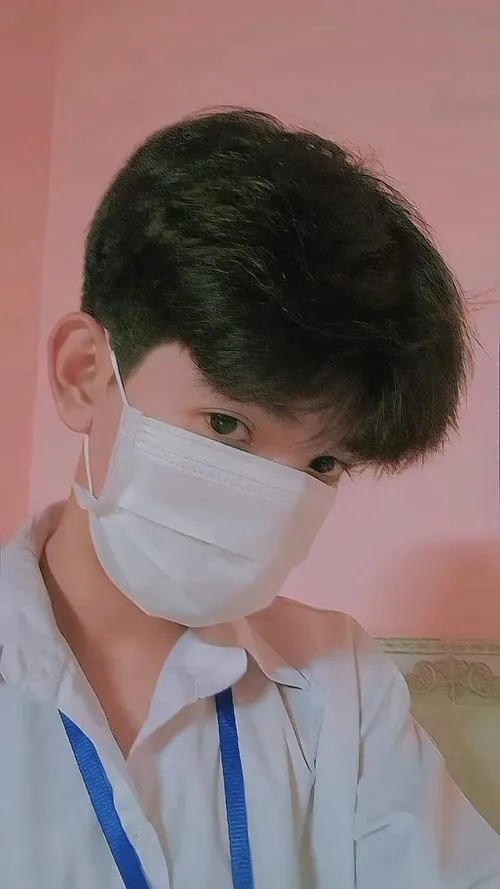 Nam sinh tóc đen mặc áo sáng màu và đeo khẩu trang trắng, nghiêng người trước nền tường nâu trong khung ảnh cận.